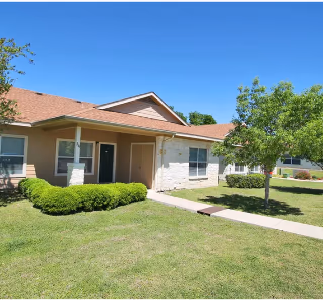 Single-story residential building with a front porch, walkway, trimmed shrubs, a tree, and a grassy lawn under a clear blue sky.