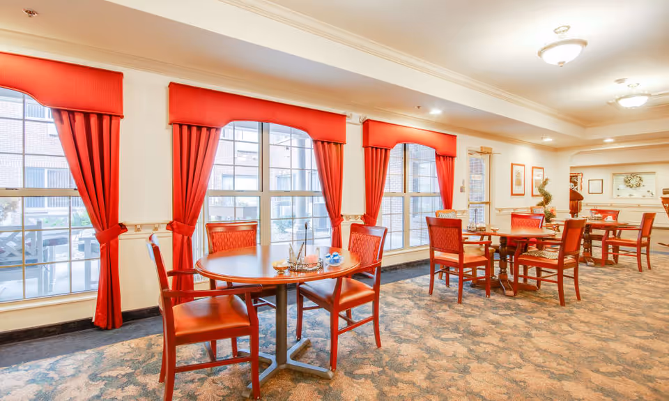 A bright dining room with several round wooden tables and red cushioned chairs. Large windows with red curtains allow natural light to fill the room. The carpet has a patterned design, and the walls are decorated with framed pictures and a wreath. Ceiling lights provide additional illumination.