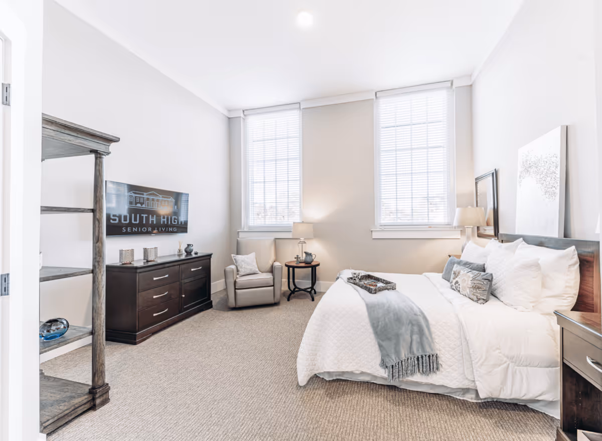A bright and neatly arranged bedroom in a senior living facility featuring a large bed with white bedding and decorative pillows, a light blue throw blanket, a wooden nightstand with a lamp, a comfortable armchair next to a small round table with a lamp, two large windows with white blinds, a dark wooden dresser with decorative items, and a wooden shelving unit. The room has light-colored walls and carpeted flooring.