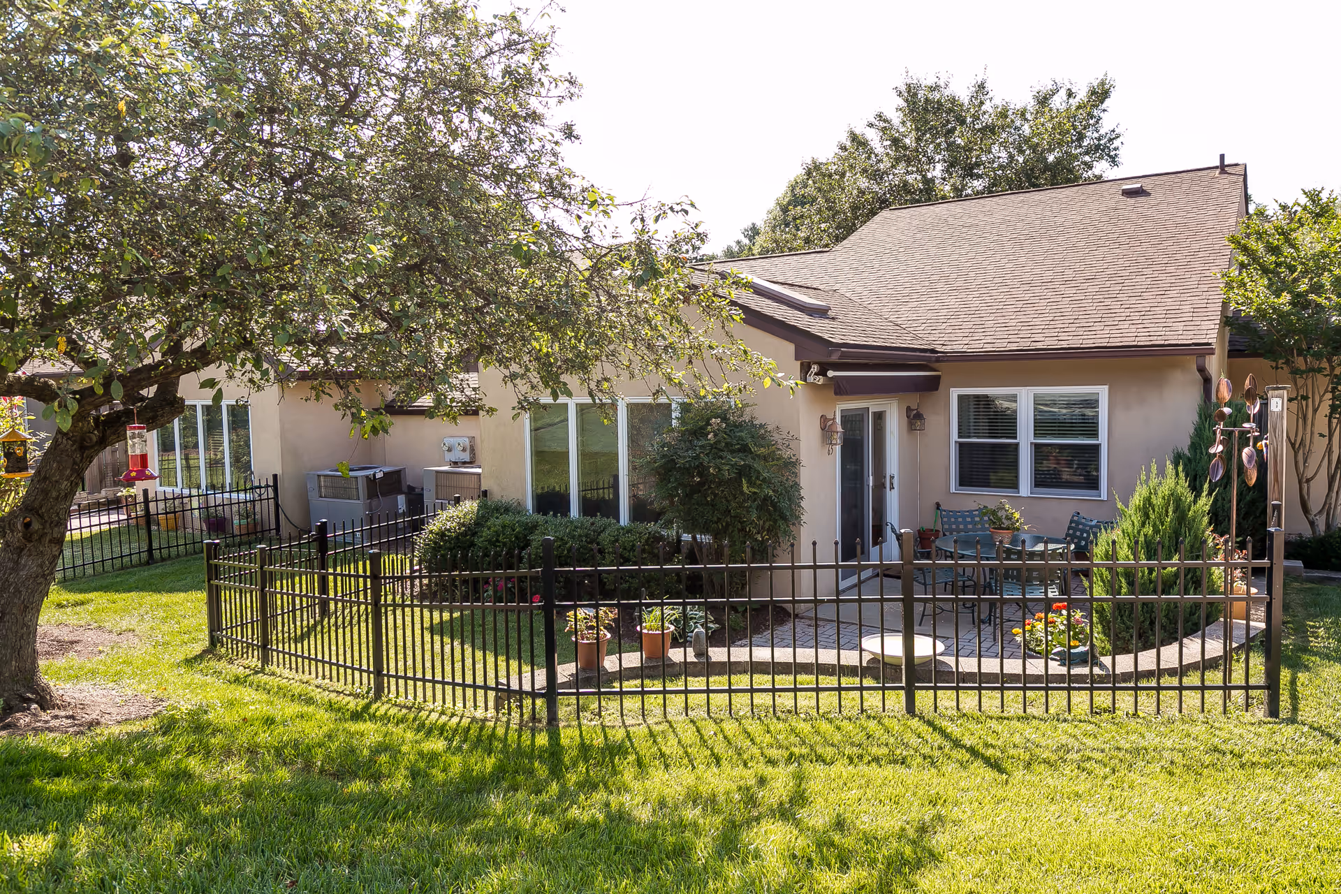 A single-story building with a fenced patio area featuring outdoor seating, potted plants, and greenery. There is a tree with a bird feeder in the foreground and well-maintained grass surrounding the area.