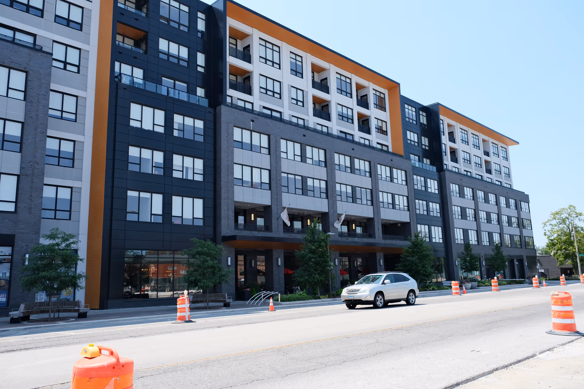 Exterior view of a modern multi-story building with large windows and balconies, situated along a street with orange construction barrels and a white SUV driving by under a clear blue sky.