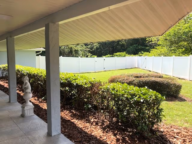 Covered porch with columns and tiled floor overlooking a fenced backyard with hedges and a grassy lawn.