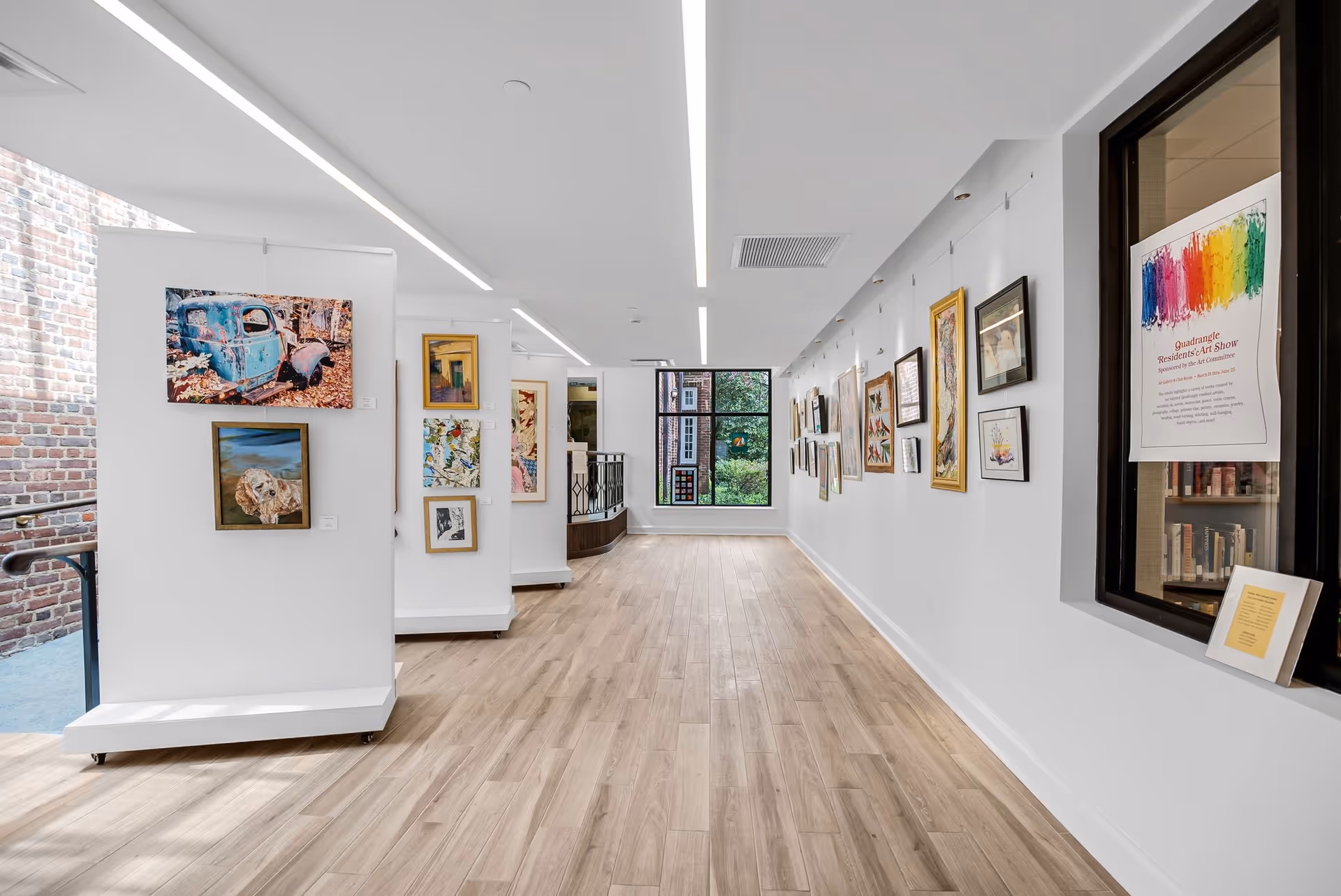 A bright, modern hallway in The Quadrangle featuring light wood flooring and white walls adorned with various framed artworks. On the left side, there are white display panels with paintings, and on the right wall, more framed art pieces are hung. A large window at the end of the hallway lets in natural light, and a colorful poster about a residents' art show is visible on the right near a window into another room.