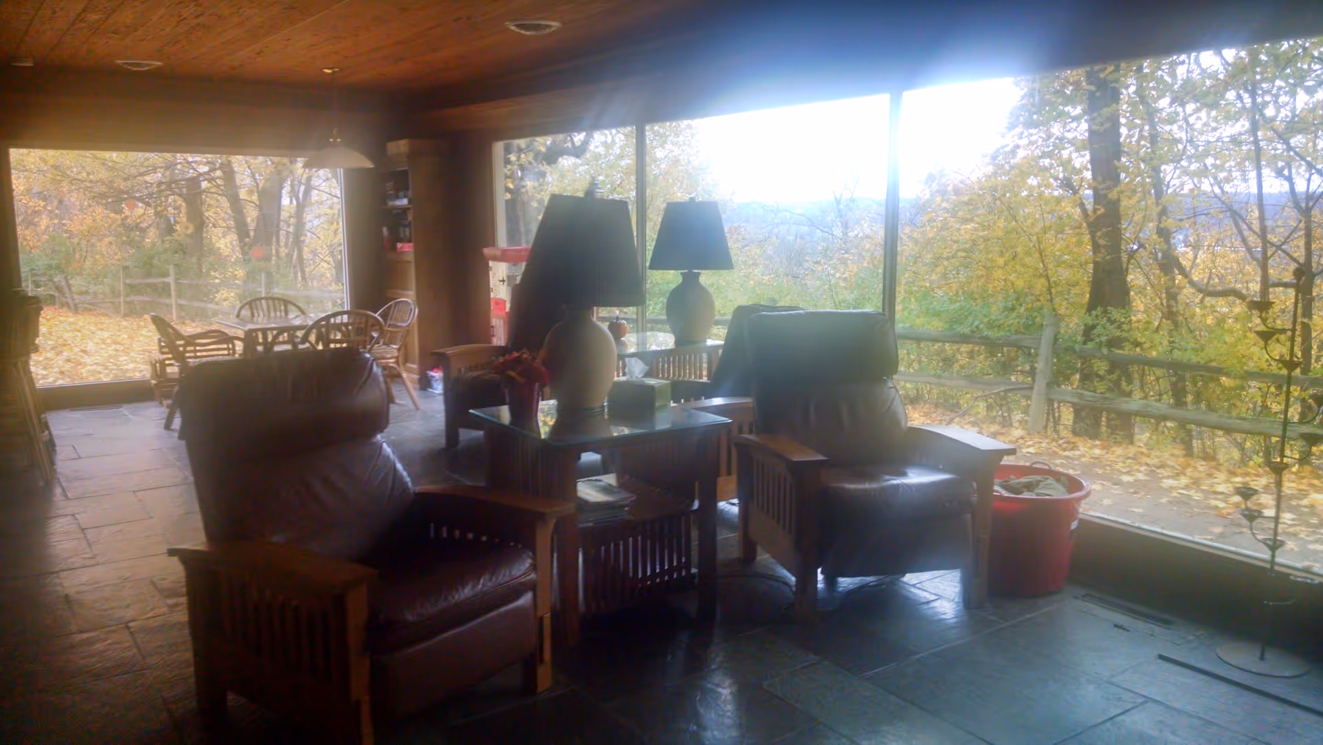 A cozy living room area with two leather armchairs and a wooden side table with a lamp on it. Large windows provide a view of an outdoor area with trees and a wooden fence, showing autumn foliage. In the background, there is a dining table with chairs near another large window.