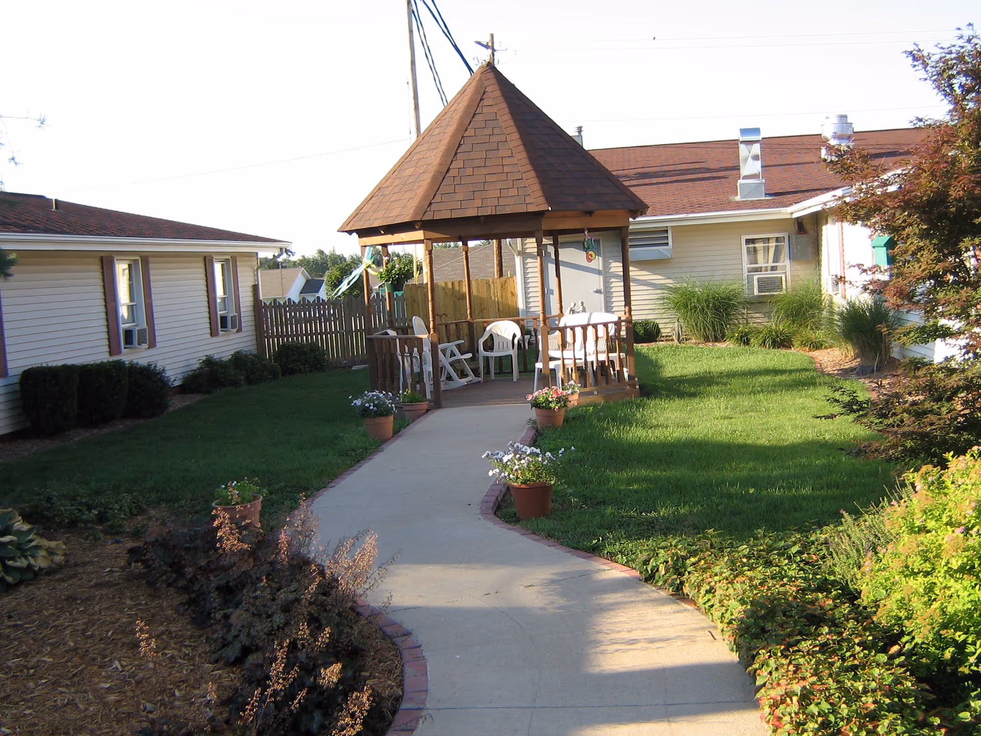 A paved walkway leading to a wooden gazebo with a brown shingled roof in a garden area. The gazebo contains white plastic chairs and a table. Surrounding the walkway and gazebo are green lawns, potted flowers, bushes, and two single-story buildings with beige siding and brown roofs.