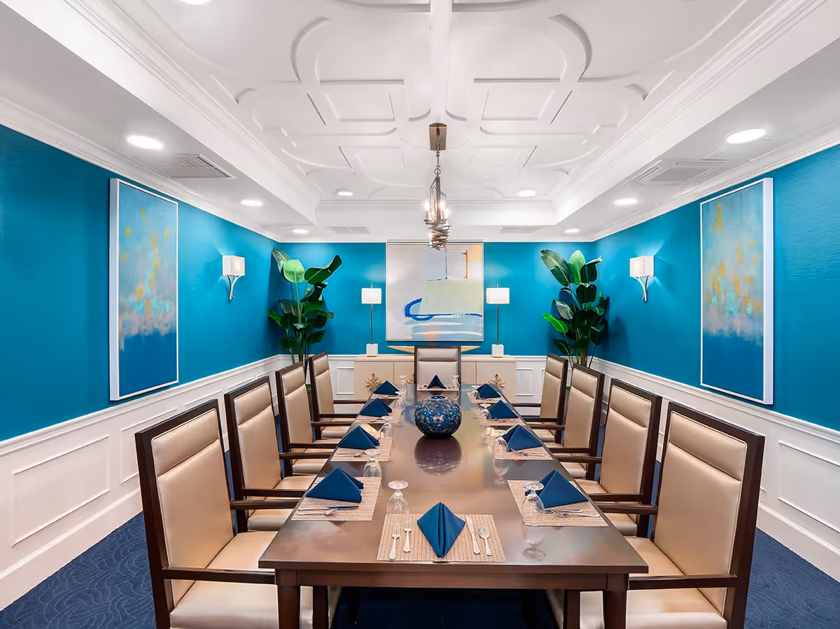 A formal dining room with a long rectangular wooden table set for twelve people. Each place setting has a blue folded napkin, a glass, and silverware on a woven placemat. The room has bright turquoise walls with white wainscoting, two large abstract paintings, and two tall green plants in the corners. The ceiling features decorative molding and a modern chandelier hanging above the table.