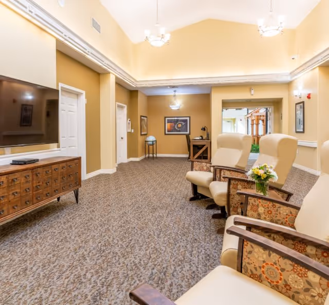 Bright senior living common room with upholstered armchairs facing a wall-mounted TV, patterned carpet, and warm yellow walls under chandelier lighting.