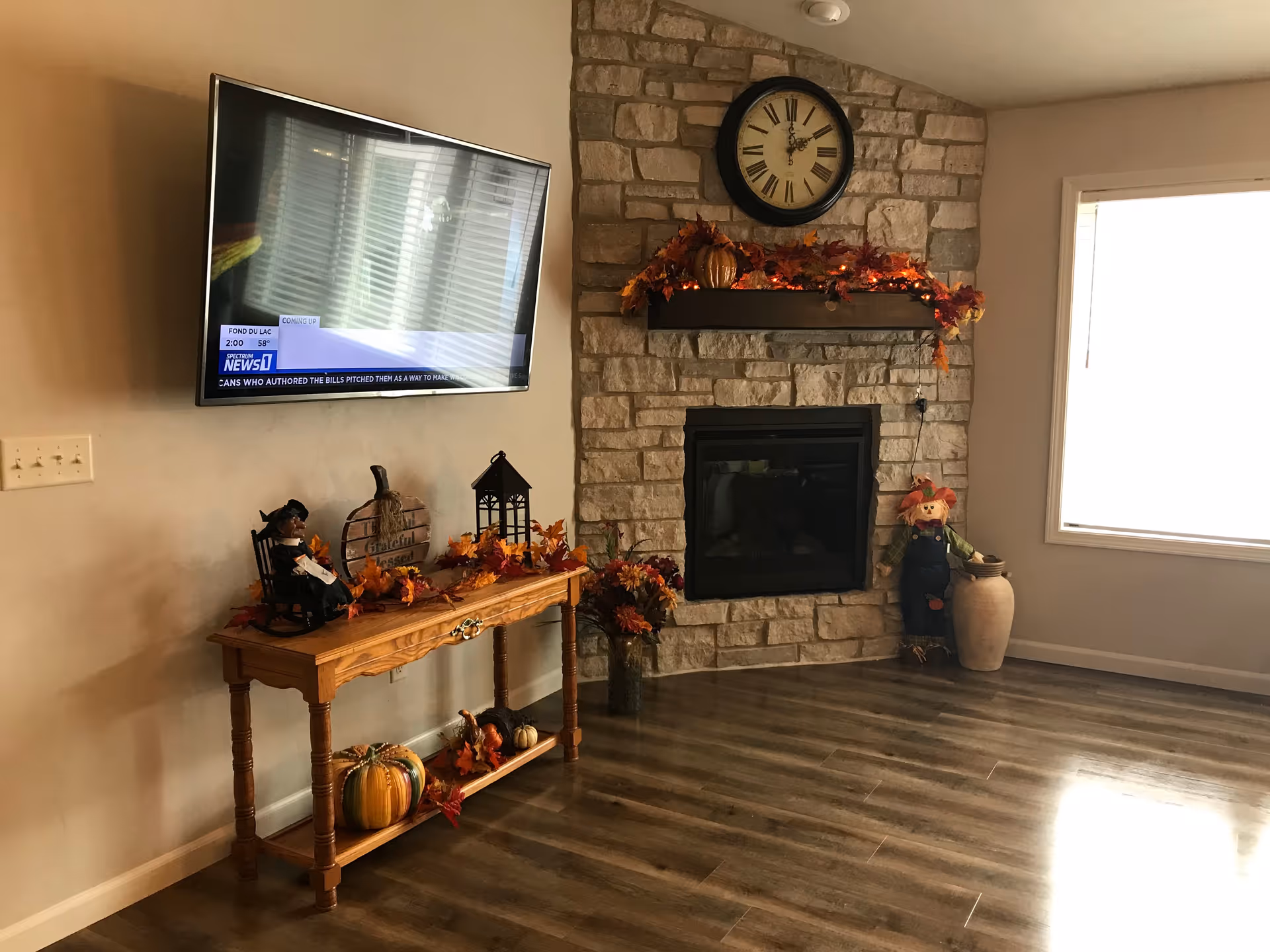 A cozy living room corner featuring a stone fireplace decorated with autumn-themed garlands and a large clock above it. To the left, a wooden console table holds fall decorations including pumpkins, a small rocking chair figurine, and a lantern. A flat-screen TV is mounted on the wall above the table. The room has wood flooring and a large window letting in natural light.