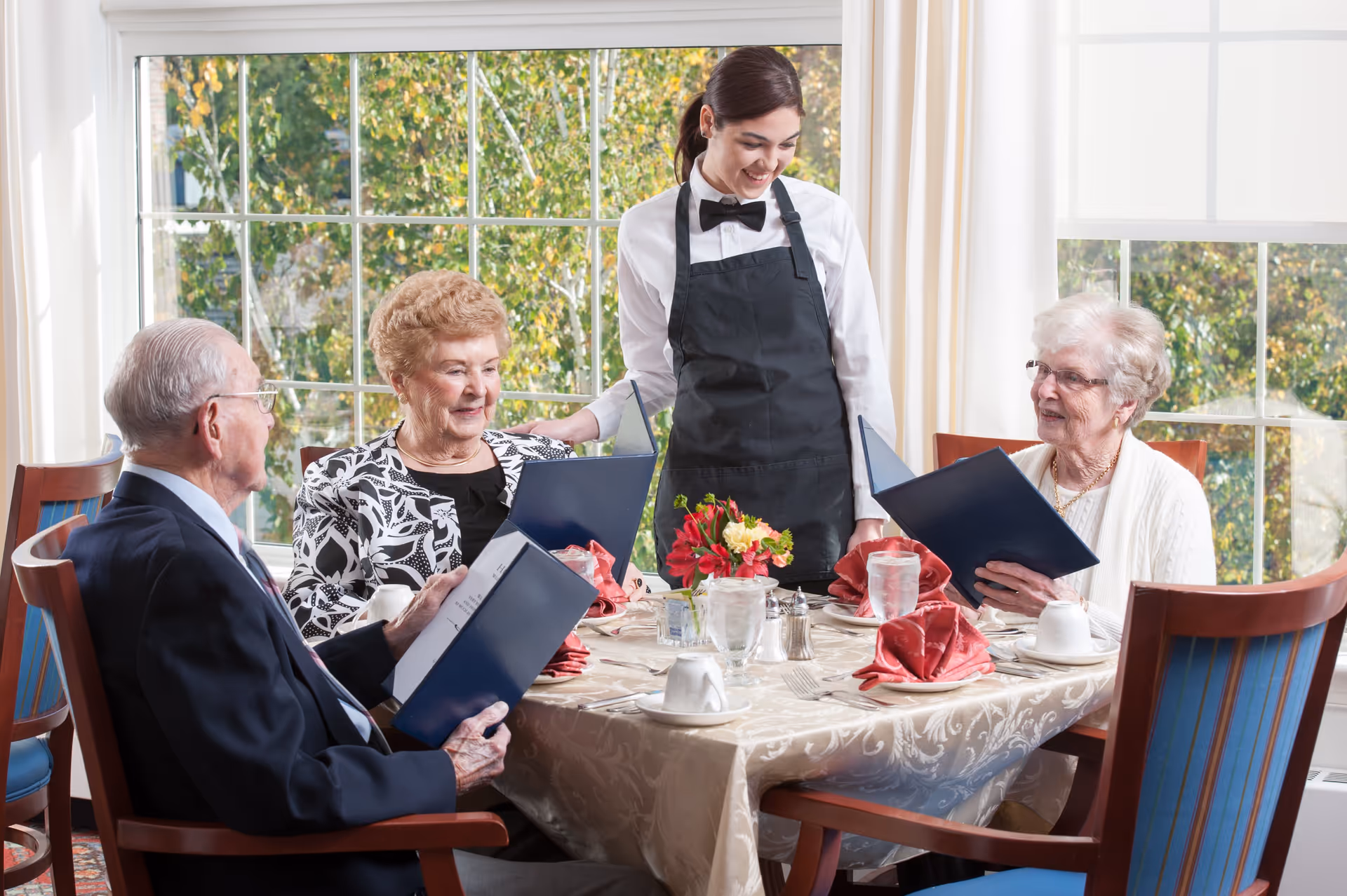 A waitress in a black apron and bow tie stands at a dining table with three elderly people, two women and one man, who are looking at menus. The table is set with glasses of water, cups, red napkins, and a small flower arrangement. Large windows with white curtains show green trees outside.