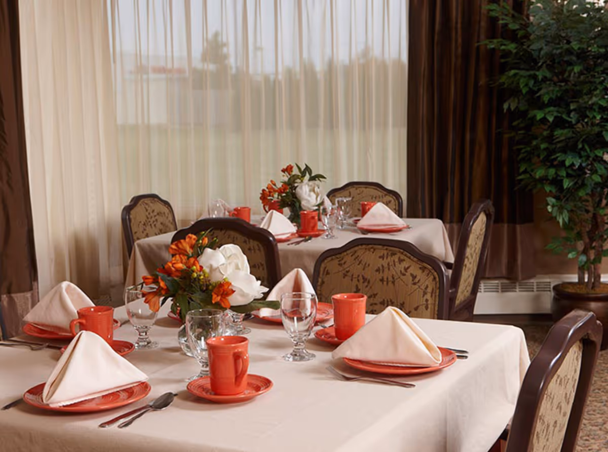 A dining room with tables set for a meal, featuring white tablecloths, red plates and mugs, folded white napkins, glassware, and floral centerpieces. The room has large windows with sheer curtains and brown drapes, and upholstered chairs with patterned fabric.