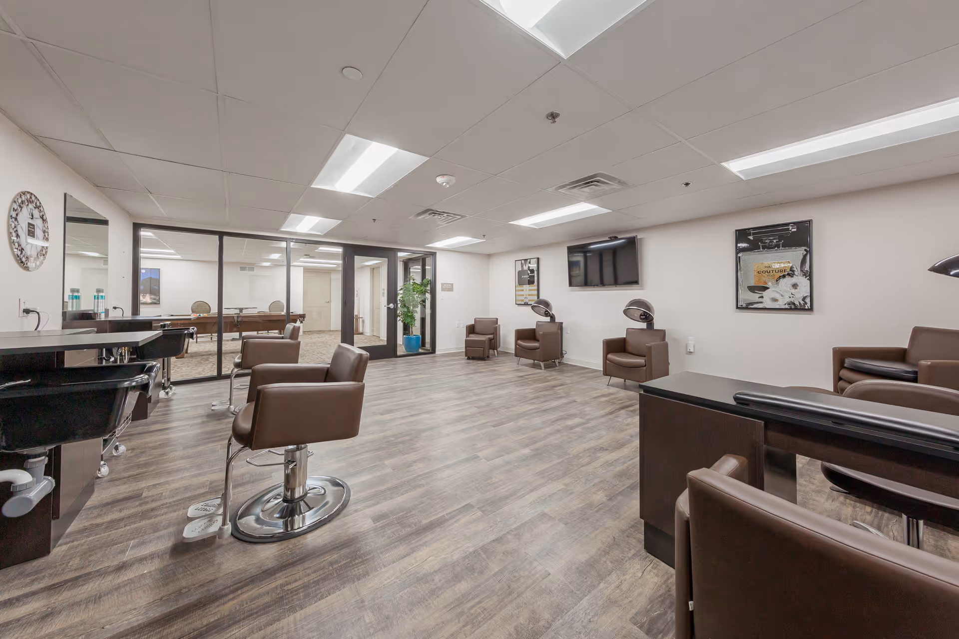 Well-lit interior salon with brown styling chairs, wash basins, hair dryers, and a wall-mounted TV.