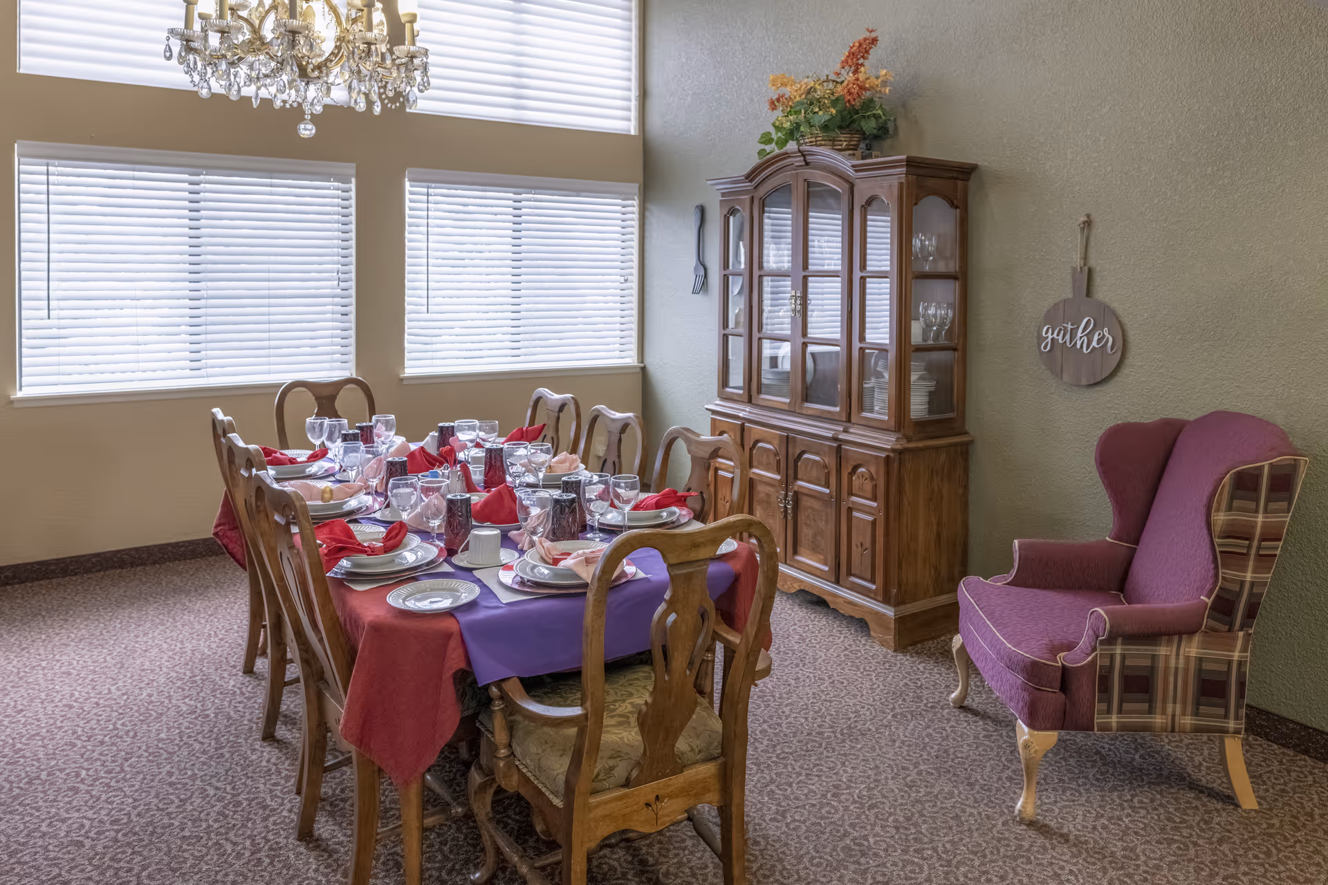 A dining room with a long wooden table set for a meal, featuring plates, glasses, and red and pink napkins. The room has large windows with blinds, a chandelier hanging from the ceiling, a wooden china cabinet filled with dishes and glassware, and a purple upholstered wingback chair with plaid sides. A decorative wall sign that says 'gather' is hanging on the wall.
