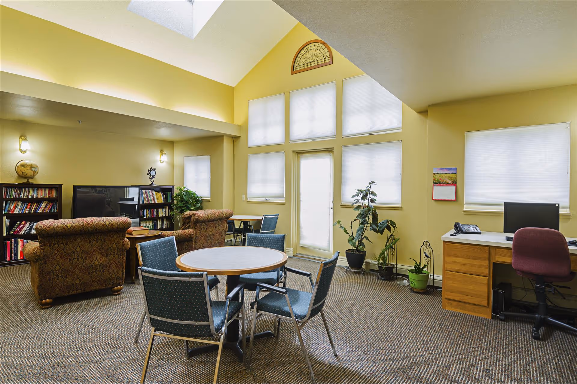 A bright communal lounge with a round table and chairs, upholstered sofas, bookshelves, a TV, a desk, and potted plants beneath tall windows.