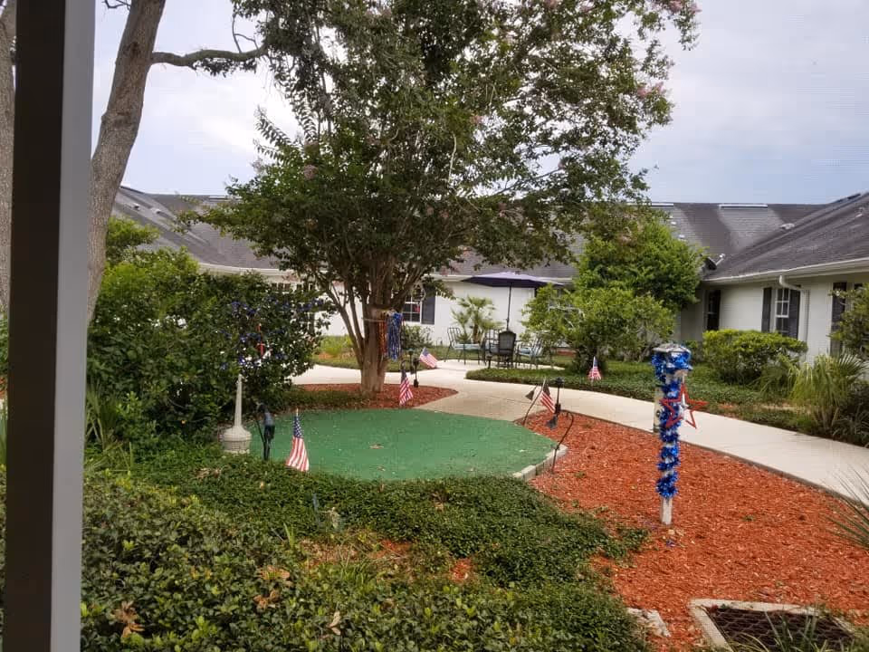 Courtyard with landscaped paths, trees, patio seating and patriotic decorations between single-story facility buildings.