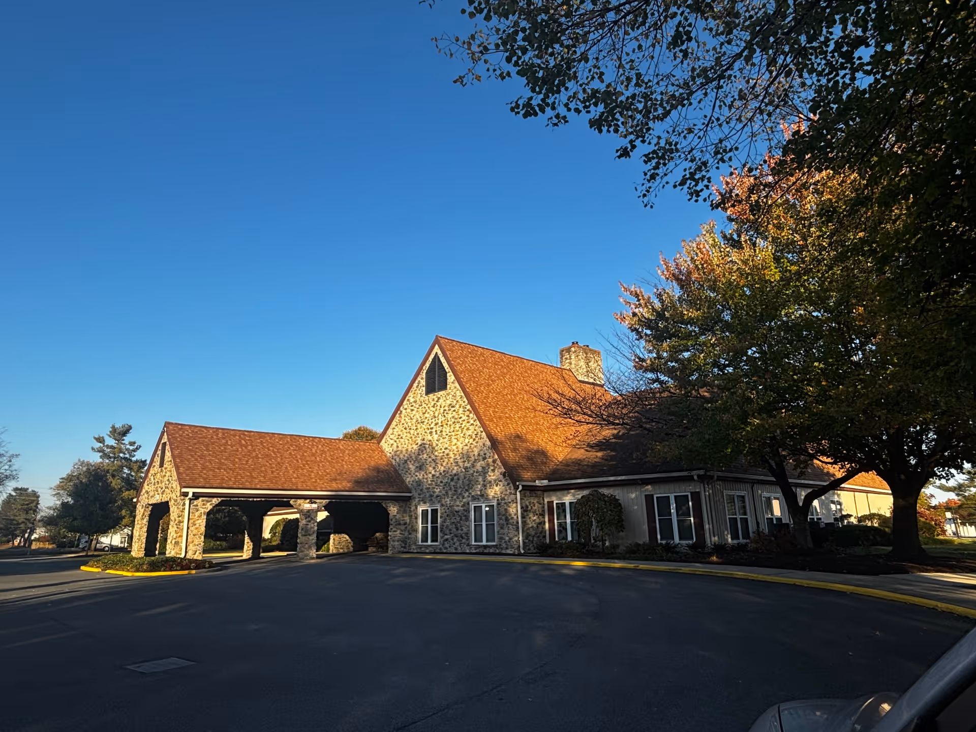 Exterior view of a stone building with a steep roof and covered entrance, surrounded by trees with autumn foliage under a clear blue sky.
