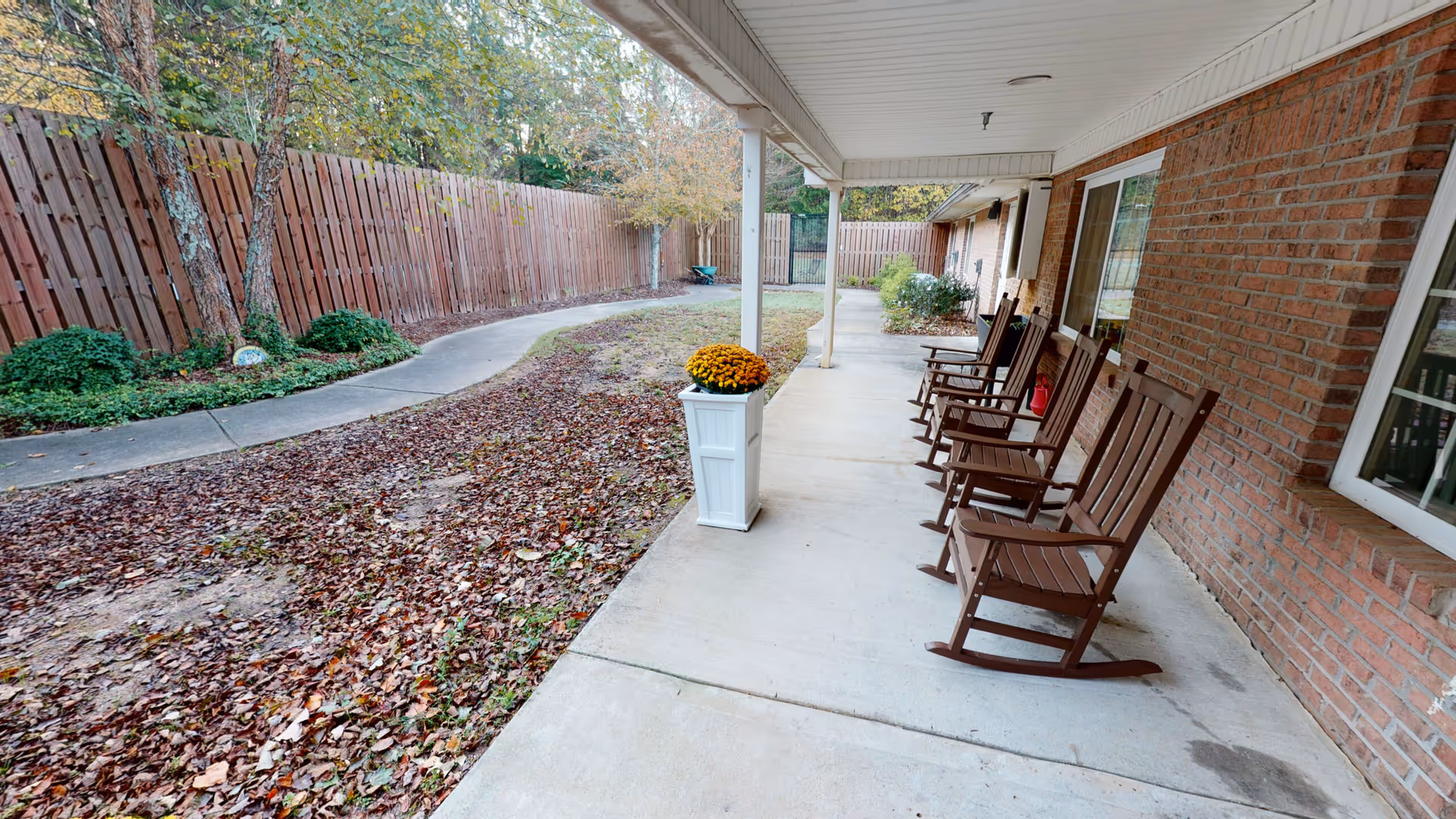 Covered outdoor patio with a row of wooden rocking chairs along a brick building, a planter of flowers, and a leaf-covered walkway beside a wooden fence.