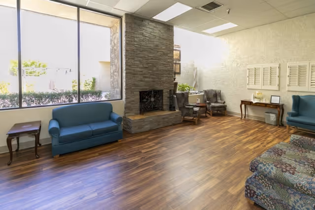 A bright common area with large windows letting in natural light, featuring a stone fireplace in the center. The room has wooden flooring, a blue sofa, patterned armchairs, a small wooden side table, and a console table with decorative items against the wall.