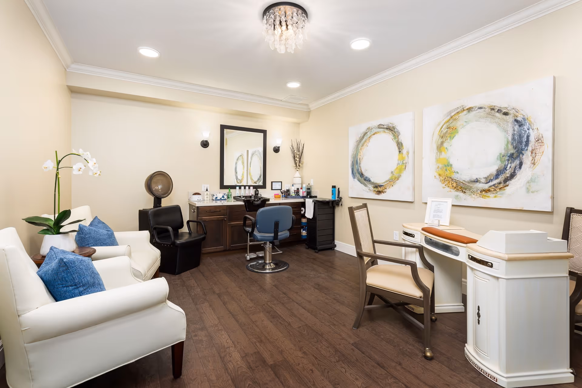 A well-lit senior living facility room with beige walls and dark wood flooring. The room features two white armchairs with blue cushions and a potted orchid on a small wooden table. There is a black salon chair with a hair dryer hood, a blue salon chair in front of a wooden vanity with a mirror, and various hair care products. On the right side, there is a white manicure table with two beige chairs and two abstract circular paintings on the wall above it. The ceiling has recessed lighting and a decorative chandelier.
