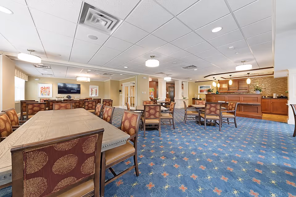 A spacious dining room with multiple wooden tables and chairs upholstered in patterned fabric. The room features a blue carpet with a floral design, beige walls, and a ceiling with recessed lighting and hanging light fixtures. There is a large flat-screen TV mounted on the far wall above a wooden sideboard, with framed artwork on either side. To the right, there is a wooden counter area with pendant lights and decorative plants.