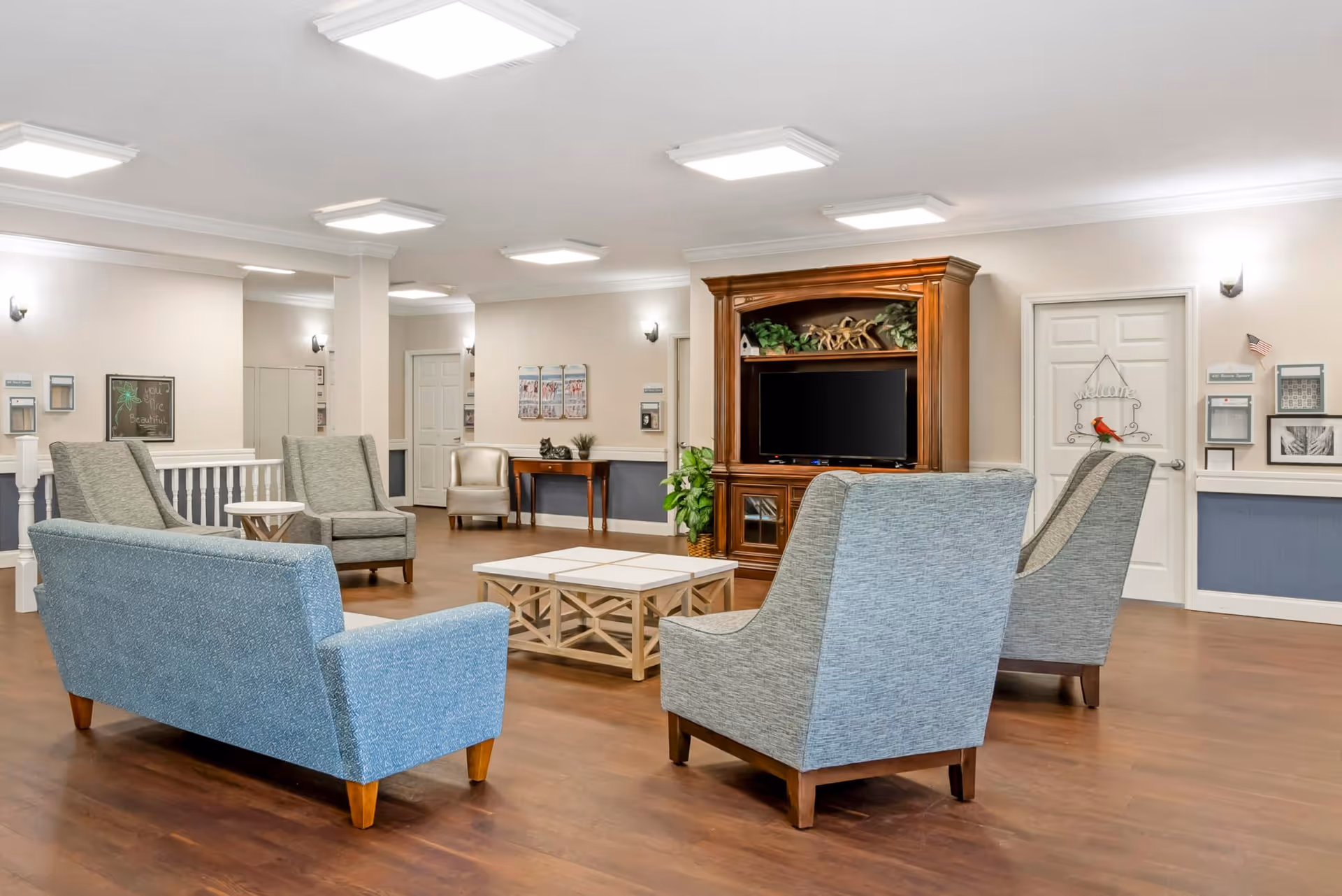 Bright communal lounge with upholstered chairs and sofas arranged around coffee tables facing a large wooden TV cabinet.