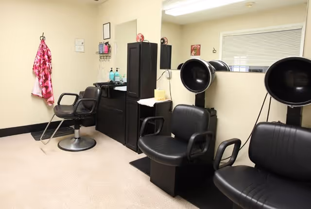 Interior of a senior living facility hair salon with three black salon chairs, two hair dryers, a black cabinet with hair care products, and a pink patterned cape hanging on the wall.