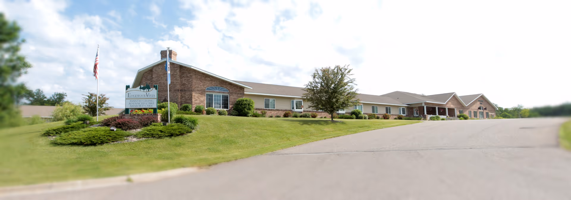 Exterior view of a single-story brick and siding building with a well-maintained lawn and landscaping. There is a sign in front that reads 'Edgewood Vista Assisted Living Advanced Care Memory Care' and two flagpoles with flags. The sky is partly cloudy.