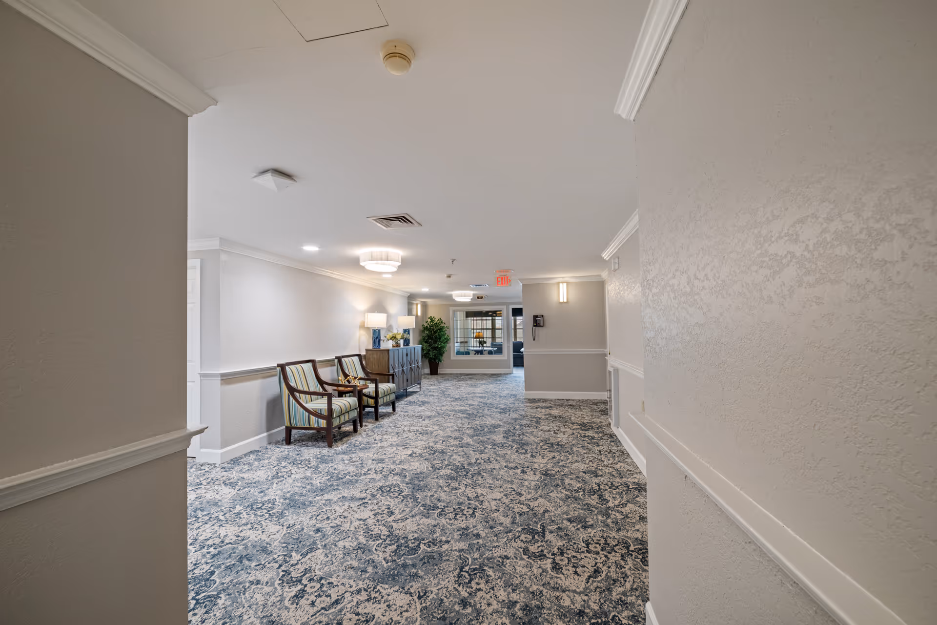 Carpeted hallway and common seating area in a senior living facility with chairs, side tables, lamps and an exit at the far end.