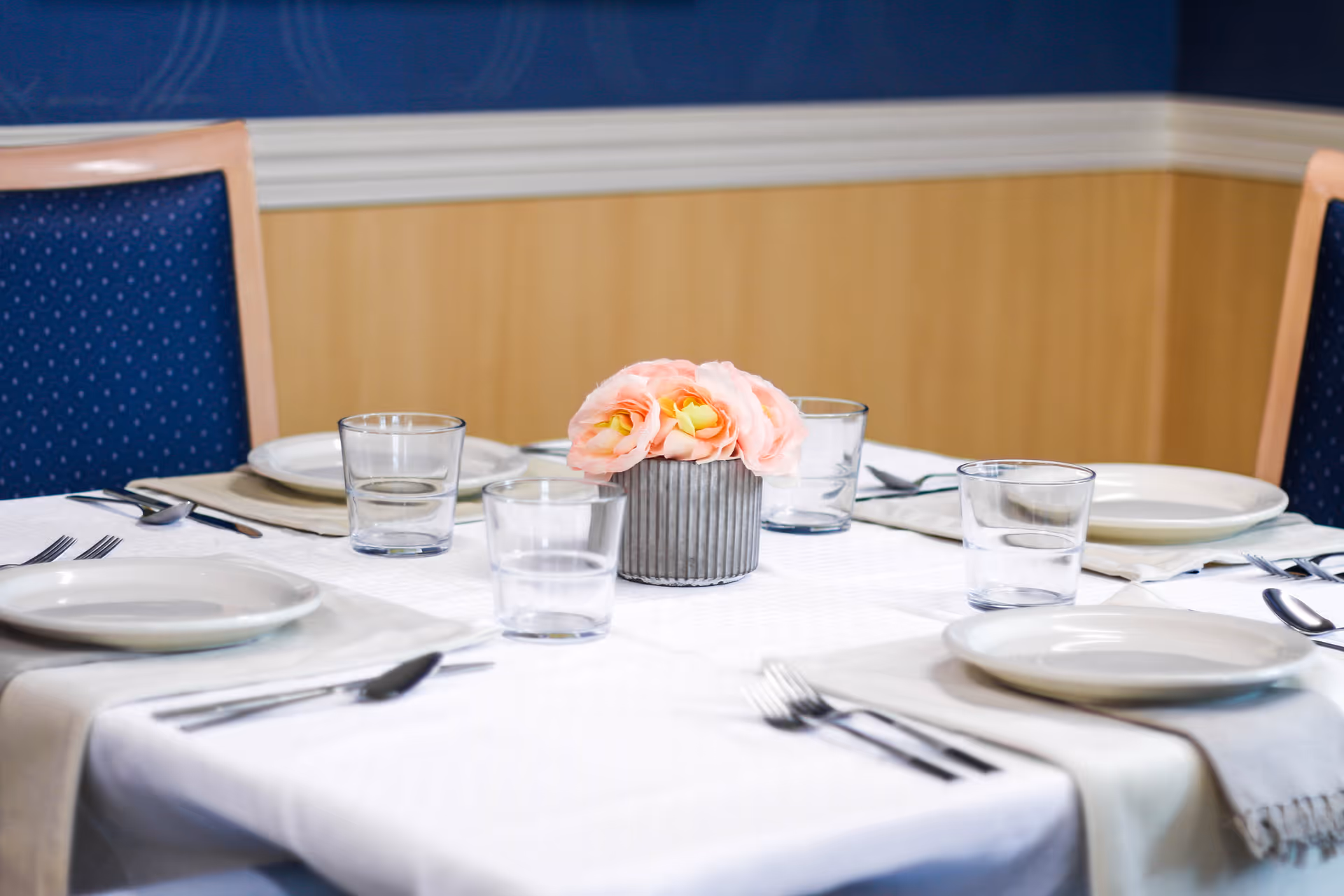 A dining table set for four with white plates, clear drinking glasses, silverware, and beige napkins. A small vase with pink flowers is placed in the center of the table. The chairs have blue upholstery with a wooden frame, and the room has a blue and beige wall background.