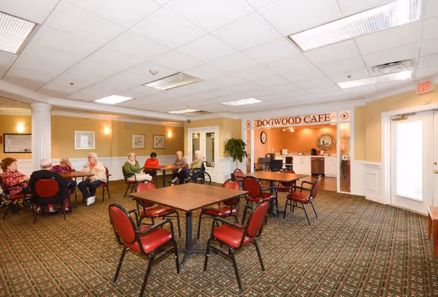 A spacious dining area with several elderly people sitting at tables and chatting. The room has patterned carpet, beige walls with white trim, and a sign above a doorway that reads 'Dogwood Cafe'. The cafe area has a counter, coffee machines, and a clock on the wall. There are red cushioned chairs around the tables and a large plant near the cafe entrance.