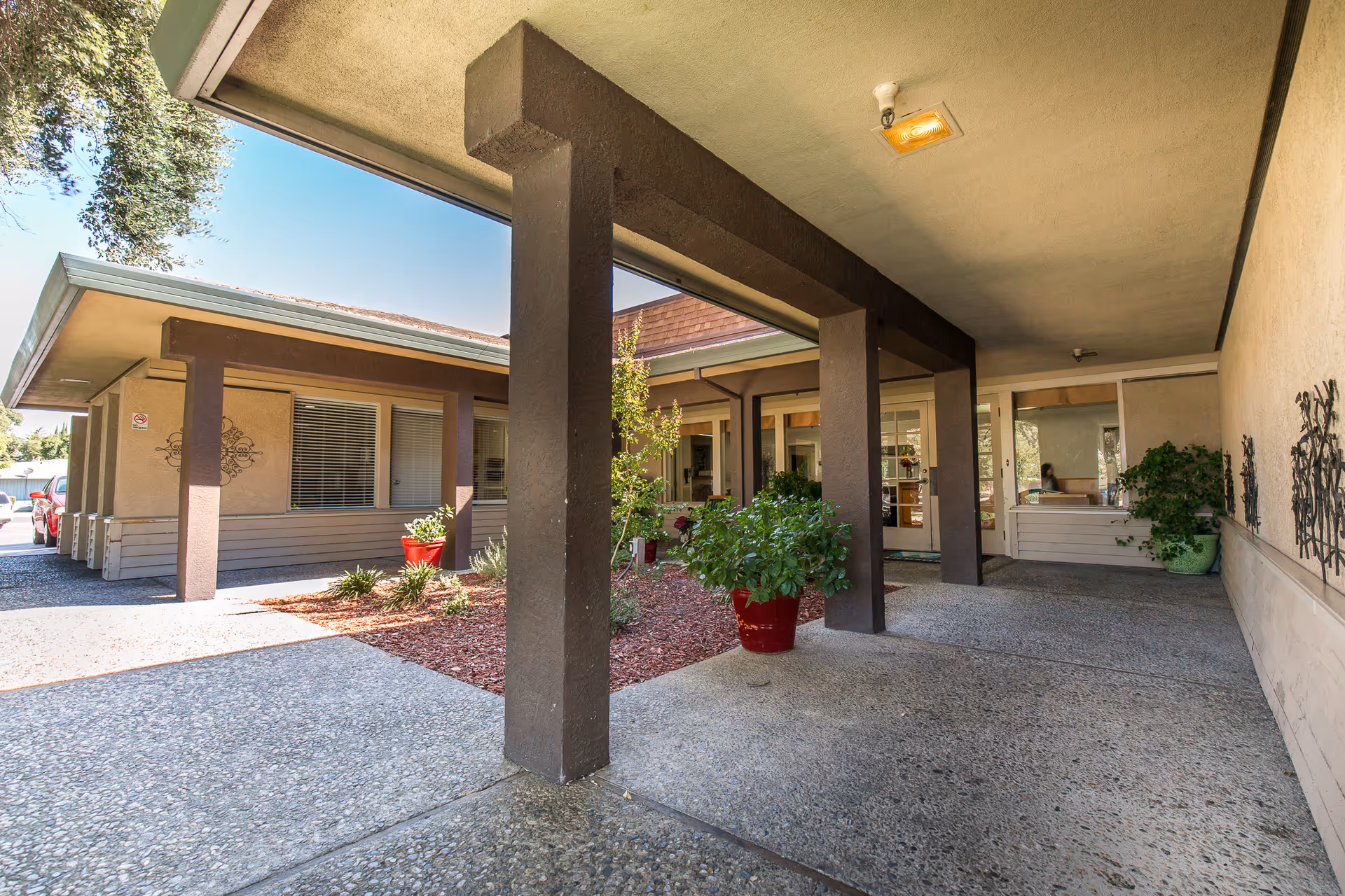 Covered entrance area of a building with brown pillars and beige walls, featuring potted plants and a pathway leading to glass doors. The sky is clear and blue, and there is some landscaping with mulch and small plants.