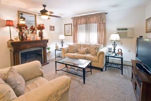 A cozy living room with beige sofas, a glass coffee table, two side tables with lamps, a wooden fireplace decorated with flowers and framed photos, a ceiling fan, a window with curtains, and a TV on a wooden stand.