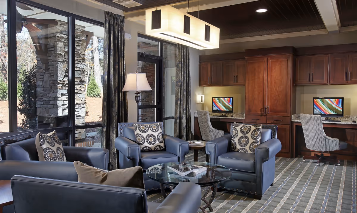 A cozy interior lounge area with four blue leather armchairs arranged around a glass coffee table. Each chair has a decorative pillow. Behind the seating area, there is a built-in wooden desk with two computer workstations and patterned chairs. Large windows with curtains provide a view of an outdoor stone pillar and greenery. A modern rectangular ceiling light fixture hangs above the seating area.