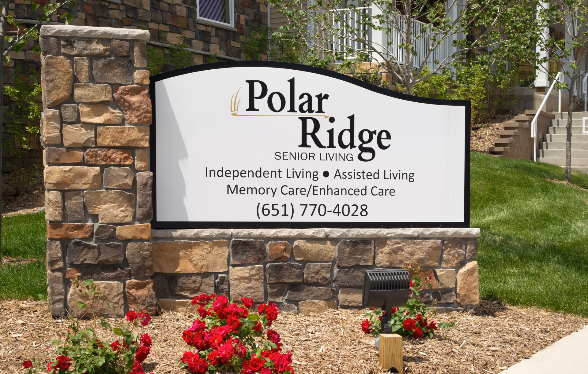 Stone-framed sign for Polar Ridge Senior Living in a landscaped front with red flowers and a nearby building entrance.