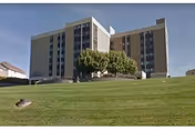 Tall beige building with a grassy lawn and trees in front near the entrance.