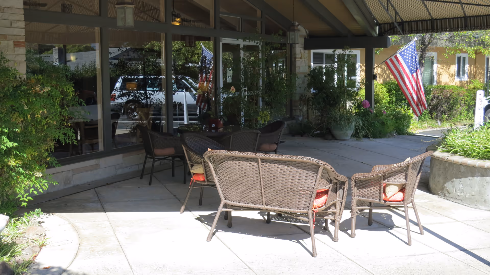 Outdoor patio area at Creekwood Care Facility with wicker chairs and a loveseat arranged around a small table. The patio is shaded by a roof and surrounded by greenery and potted plants. An American flag is displayed near the entrance of the building with large windows reflecting the outside.