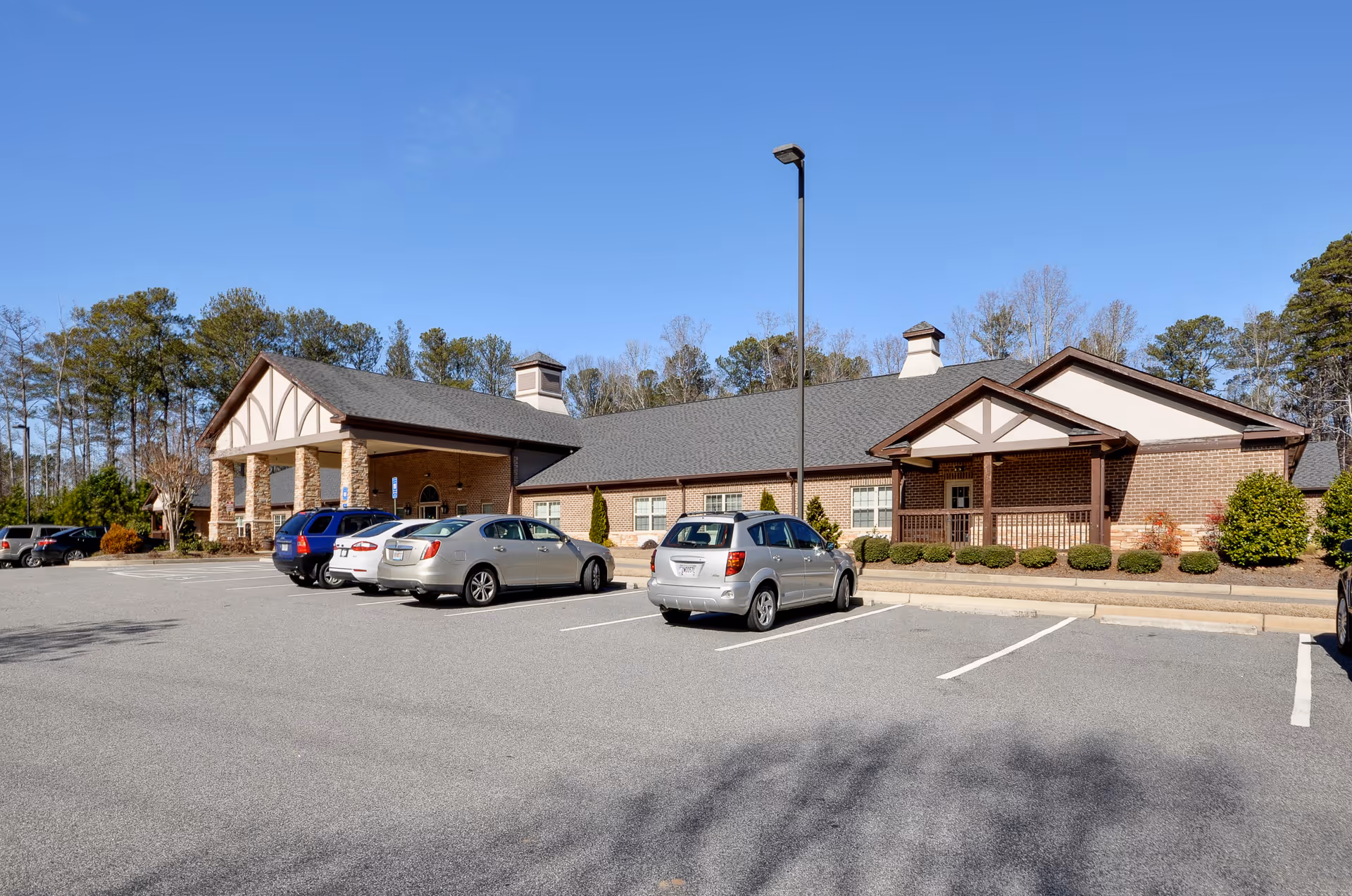 Exterior view of a single-story brick building with a gray roof and a covered entrance supported by stone columns. Several cars are parked in the parking lot in front of the building, and trees are visible in the background under a clear blue sky.