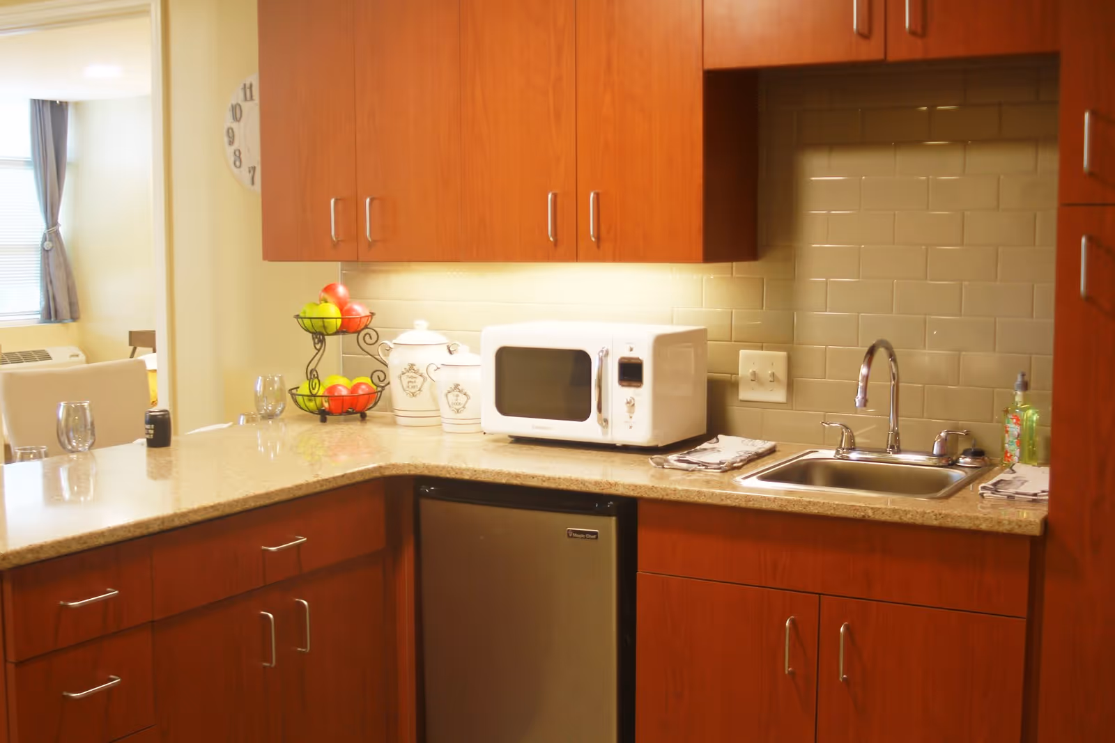 A kitchen area with wooden cabinets, a countertop with a small refrigerator underneath, a white microwave, a double-tiered fruit basket with apples, two white ceramic canisters, a stainless steel sink with a faucet, and a soap dispenser. In the background, there is a dining area with chairs and a window with curtains.
