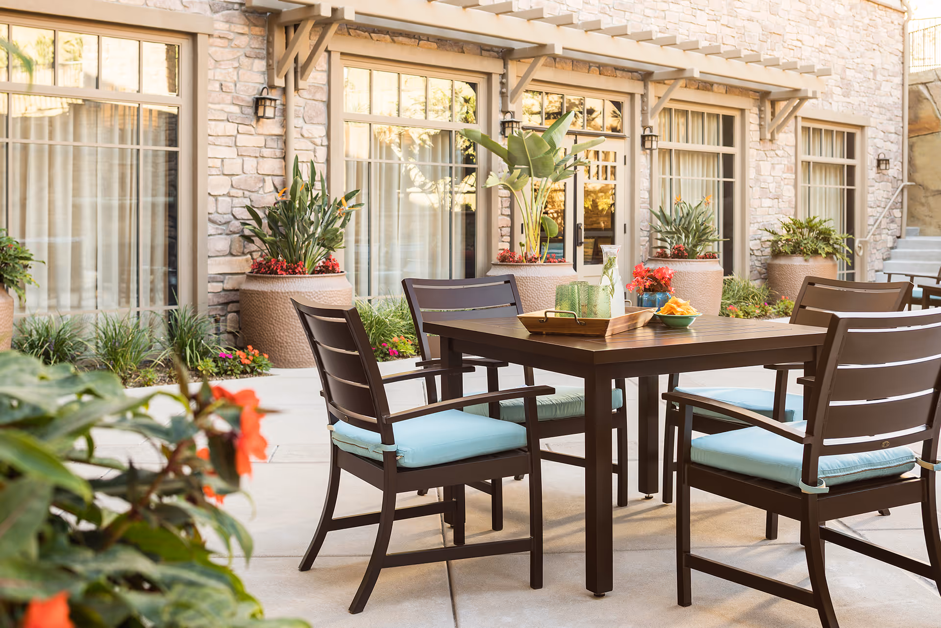 Outdoor patio with a wooden dining table and four chairs with light-blue cushions, potted plants, and stone building windows in the background.
