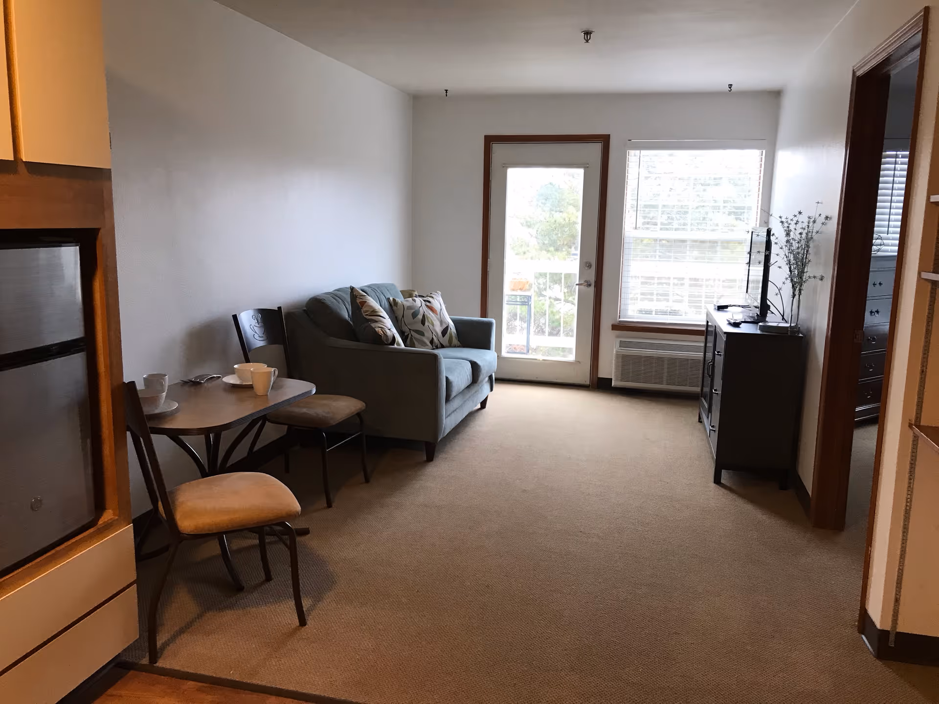 A small living area in a senior living facility with a gray loveseat adorned with patterned cushions, a small dining table with two chairs and cups on it, a TV on a dark wooden cabinet, and a door and window letting in natural light. The room has beige carpeting and white walls with wooden trim.