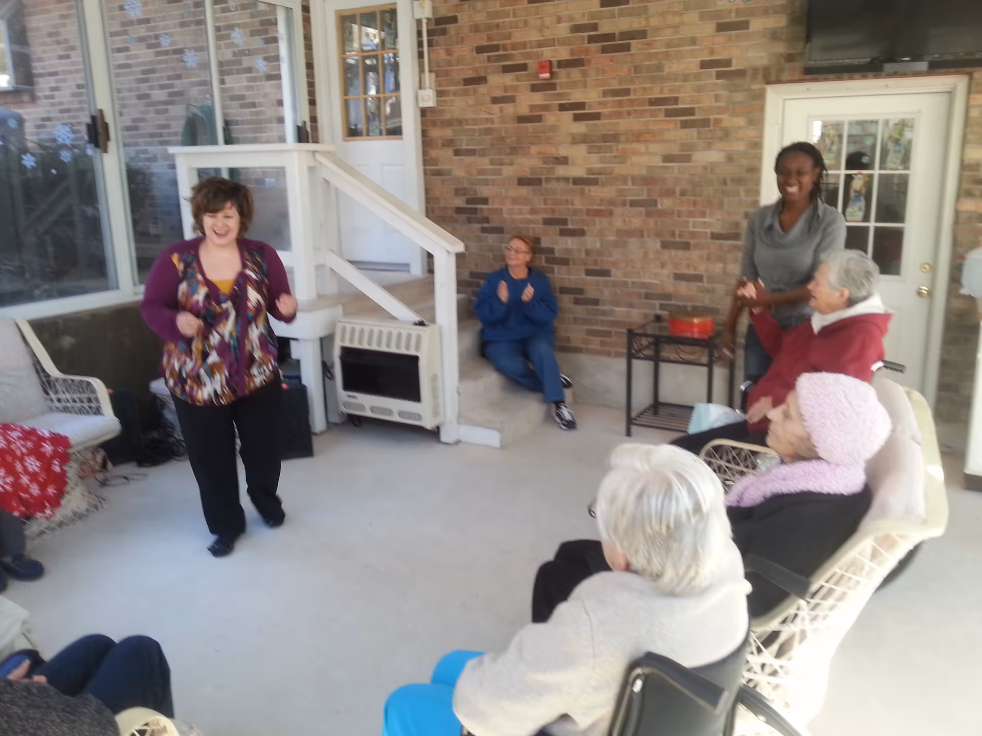 A group of elderly people and two caregivers gathered in a sunroom or enclosed patio area with brick walls and large windows. Some elderly individuals are seated in chairs, while one caregiver stands and smiles, and another caregiver is seated on a step clapping. The atmosphere appears warm and social.