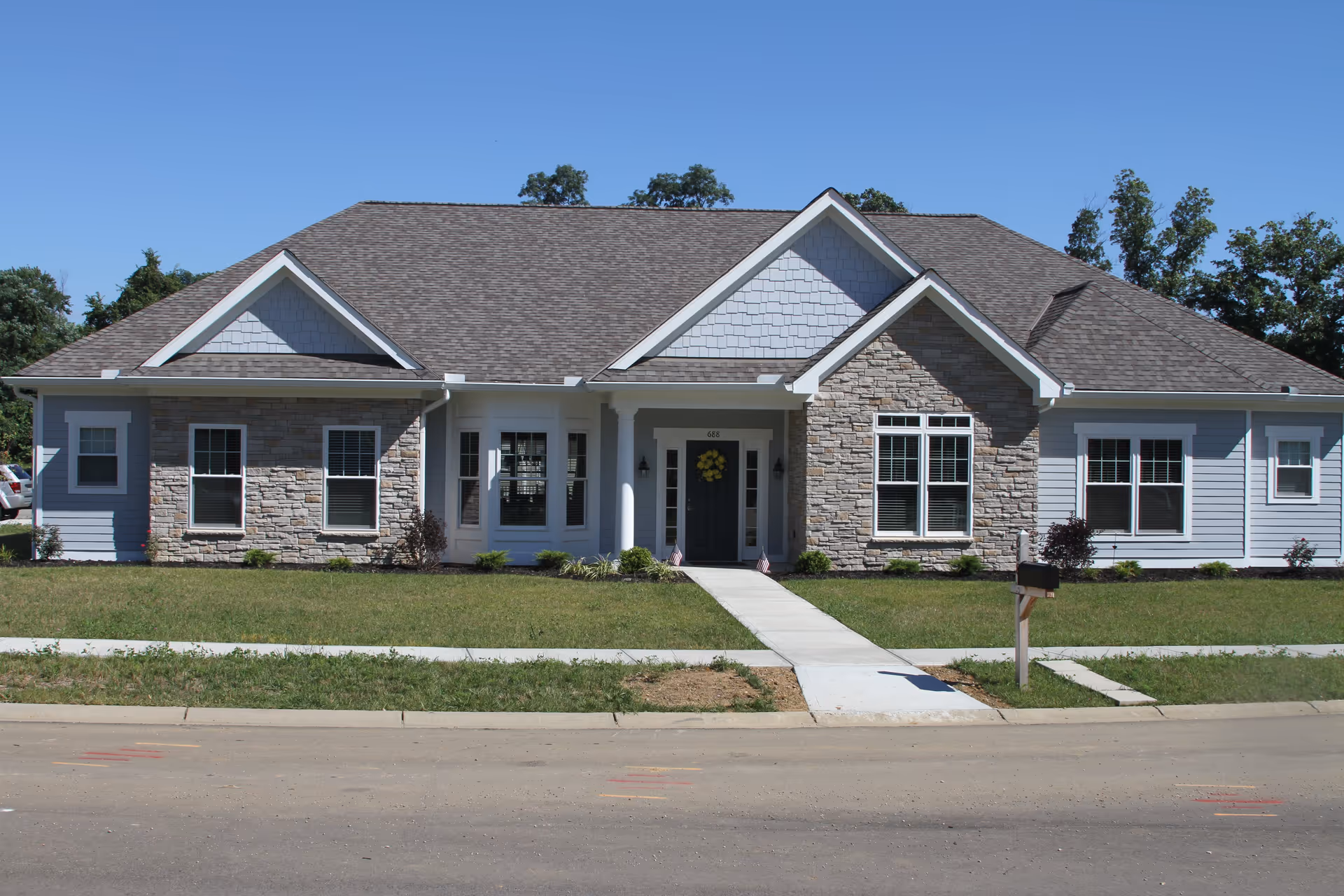 Front exterior view of a single-story senior living facility building with a gray shingled roof, stone and light blue siding, multiple windows, a central entrance with a small porch, and a walkway leading to the street.