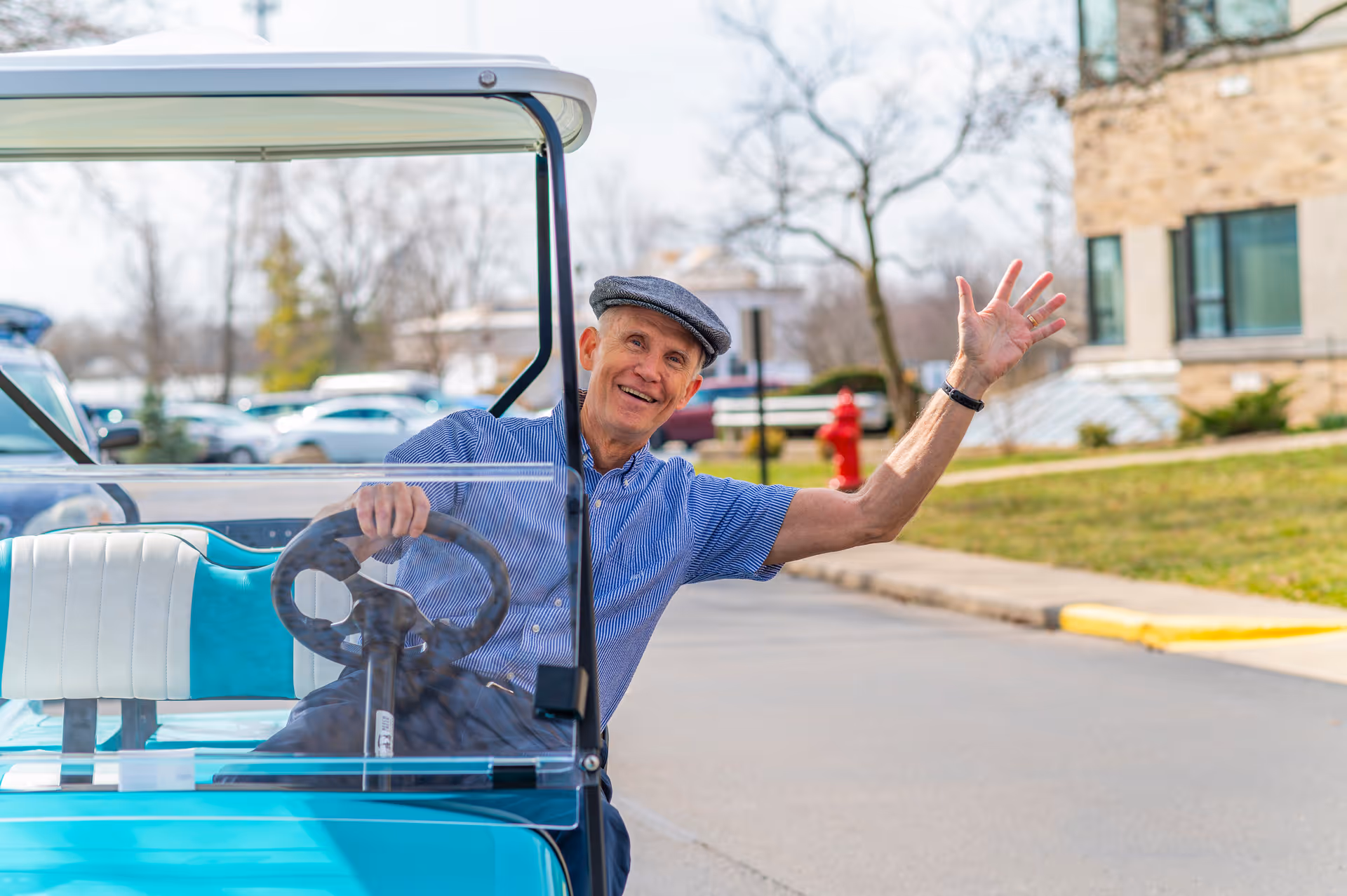 An elderly man wearing a blue shirt and a flat cap is sitting in a turquoise and white golf cart, smiling and waving with his right hand. The background shows a paved road, some parked cars, leafless trees, and a building with large windows.