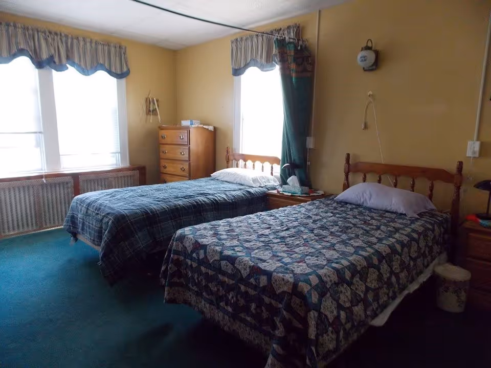 A bedroom with two single beds, each with patterned bedspreads. There is a wooden dresser between two windows with curtains and valances. A nightstand with tissues and other items is positioned between the beds. The walls are painted yellow and the floor is carpeted in green.