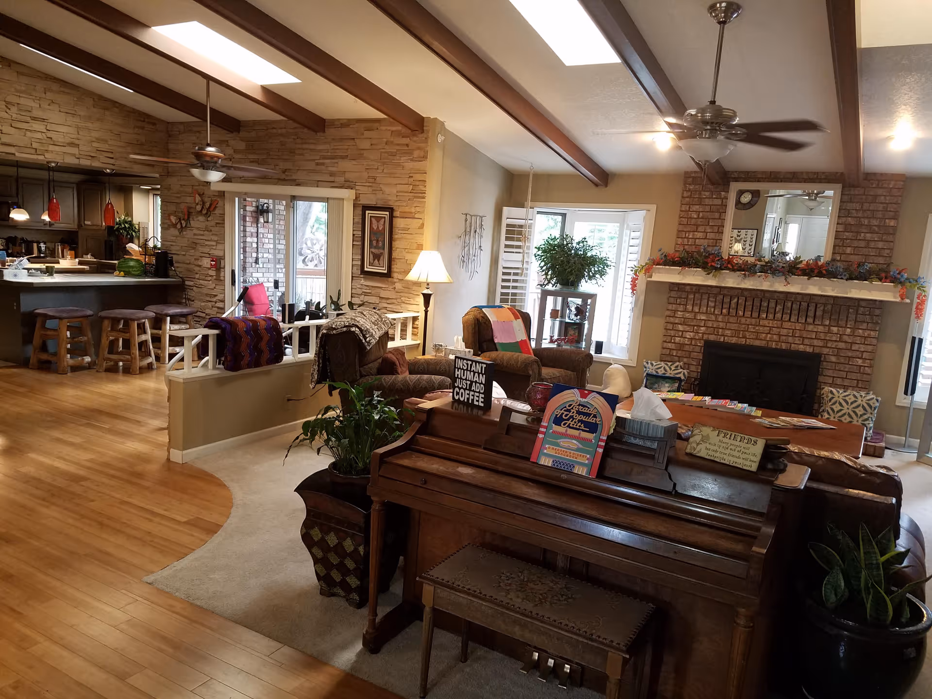 A cozy living room with a piano in the foreground, armchairs with colorful blankets, a brick fireplace decorated with garlands, and a kitchen area with bar stools in the background. The room has wooden beams on the ceiling, hardwood floors, and large windows letting in natural light.