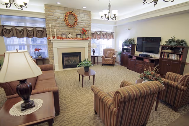 A cozy living room with a stone fireplace decorated with a wreath and autumn-themed garland. The room features a brown sofa, two striped armchairs, a wooden coffee table with a plant, a side table with a lamp, and a large flat-screen TV on a wooden entertainment center. The windows have brown valances, and the carpet has a subtle pattern.