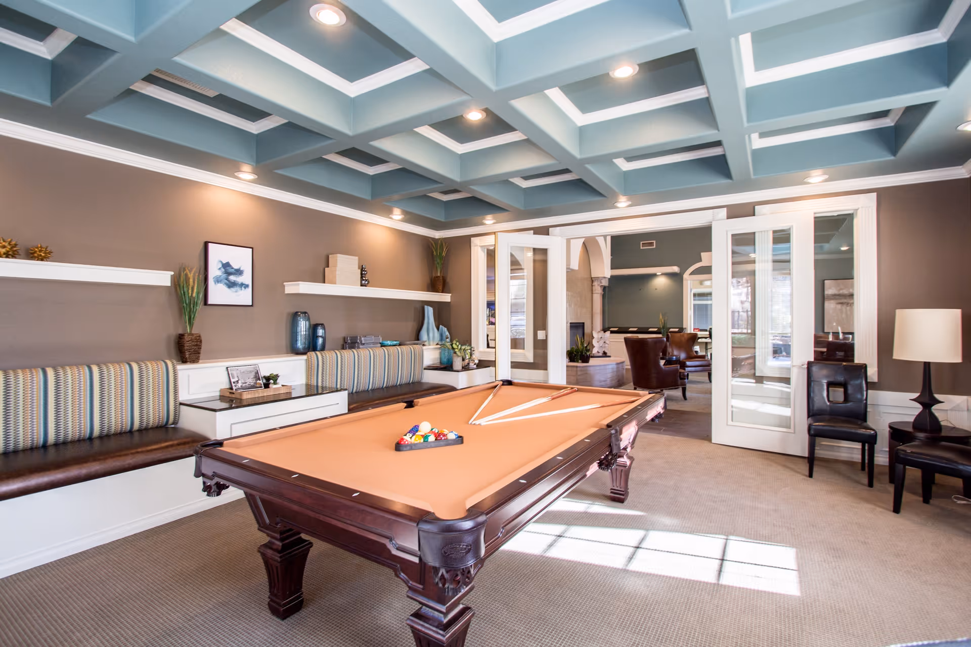 A well-lit recreational room featuring a pool table with cues and balls arranged for a game. The room has a coffered ceiling with recessed lighting, a built-in bench with striped cushions along one wall, decorative shelves with vases and plants, and glass double doors leading to another seating area with chairs and a fireplace.