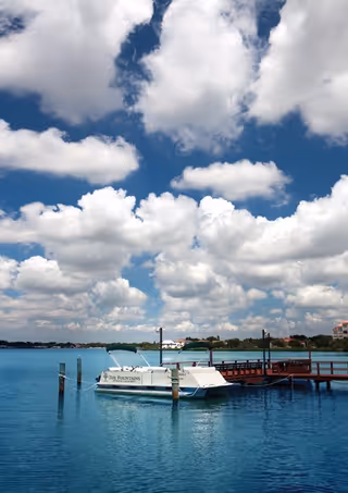 A white boat named 'The Fountains' docked at a wooden pier on a calm blue body of water under a sky filled with large, fluffy white clouds.