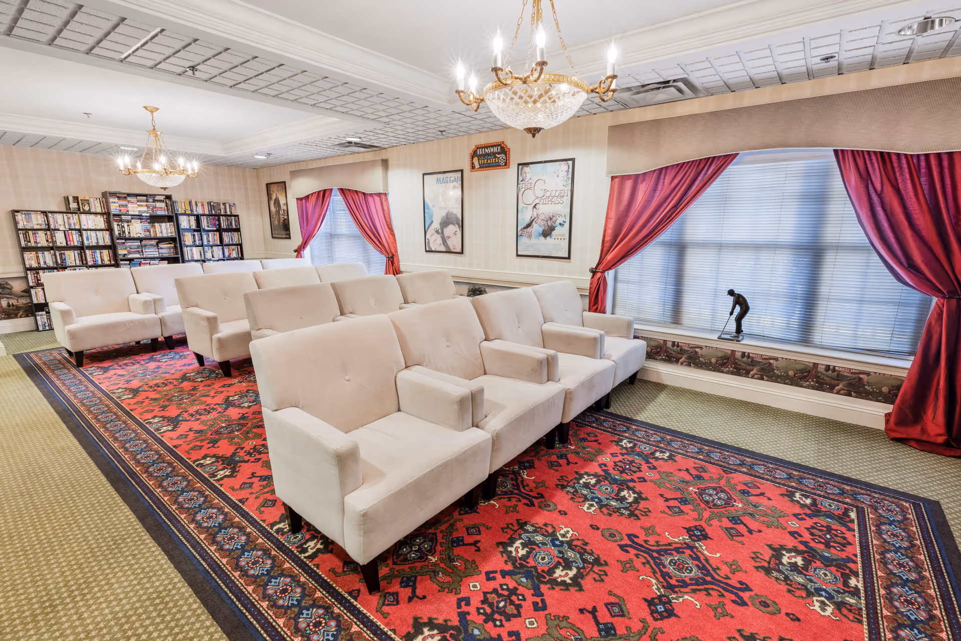 A cozy common area with rows of beige armchairs arranged on a large red patterned rug. The room features two large windows with red curtains, a bookshelf filled with books, and framed movie posters on the wall. Two chandeliers hang from the ceiling, providing warm lighting.