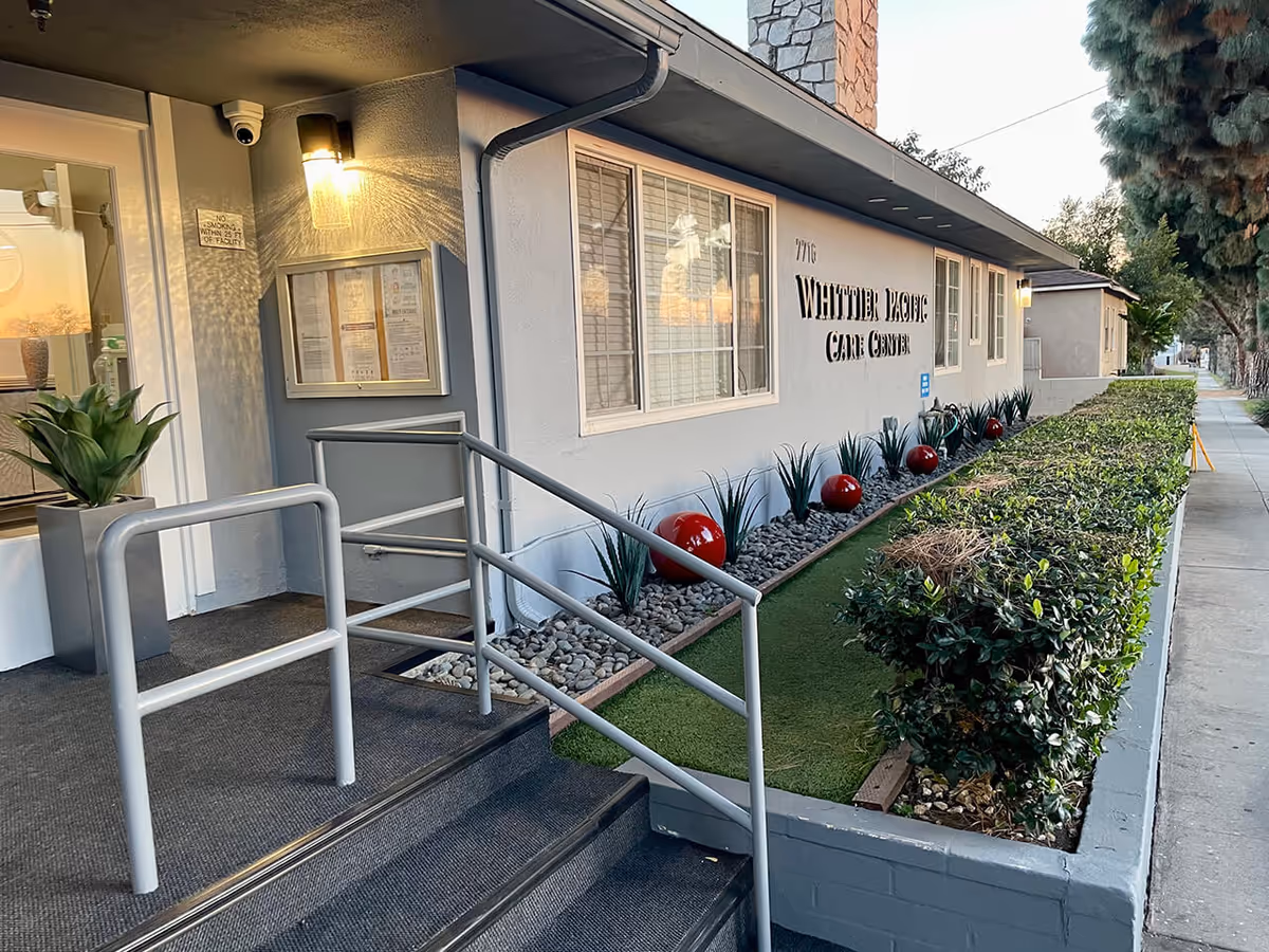 Front entrance of Whittier Pacific Care Center showing steps and handrails, a landscaped planter with red decorative spheres, and the building facade.