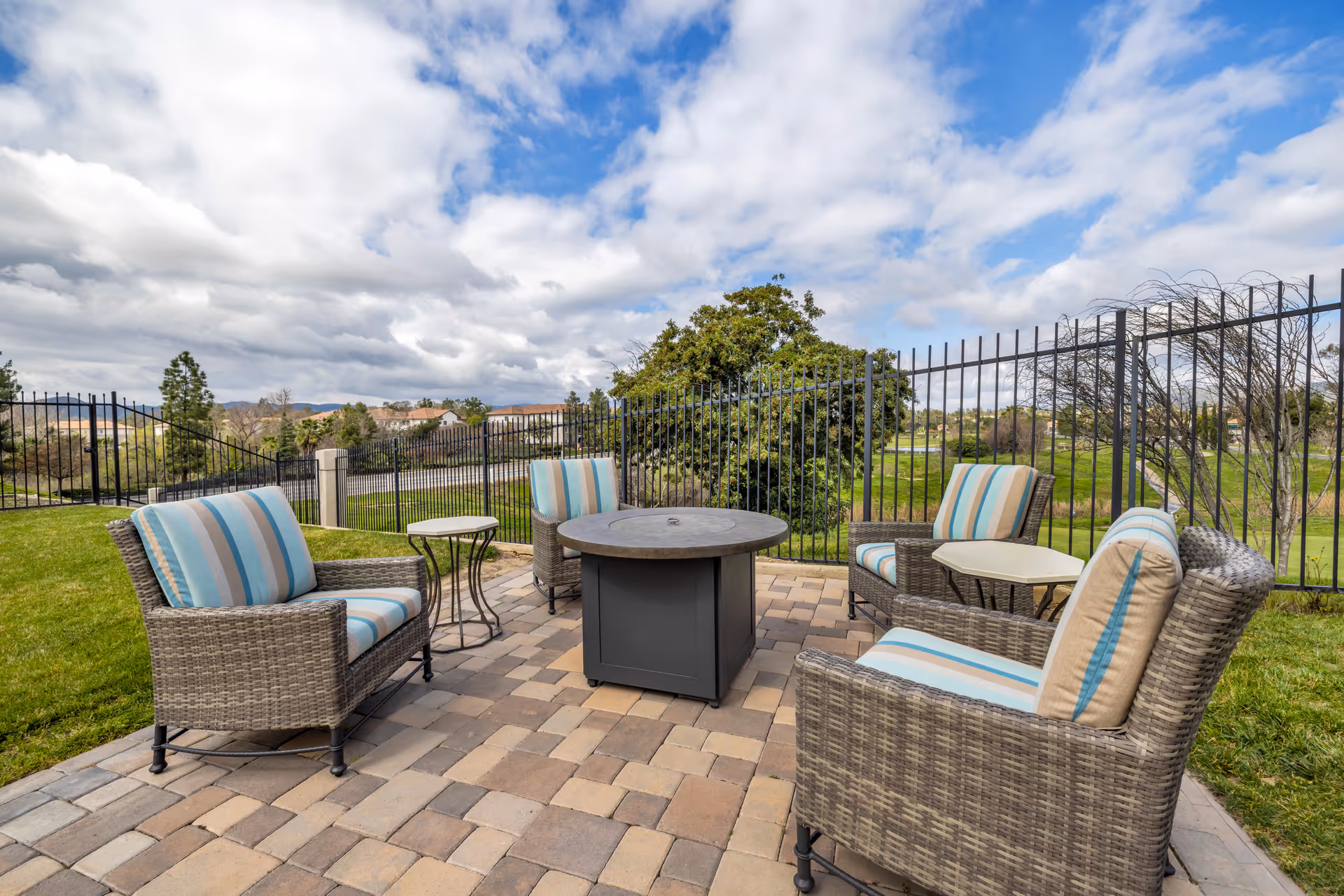 Outdoor patio area with four wicker armchairs featuring striped cushions arranged around a central fire pit table on a paved surface, surrounded by grass and enclosed by a black metal fence under a partly cloudy sky.
